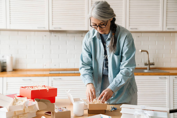 Mature grey woman in eyeglasses while wrapping present at home - Foto, imagen