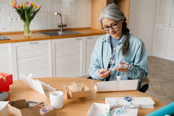 Mature grey woman smiling while wrapping present in kitchen at home - Φωτογραφία, εικόνα