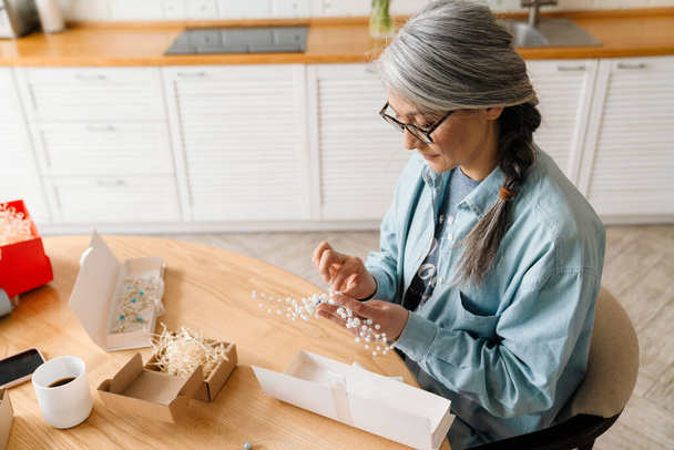 Mature grey woman in eyeglasses wrapping present at home - Foto, Imagem