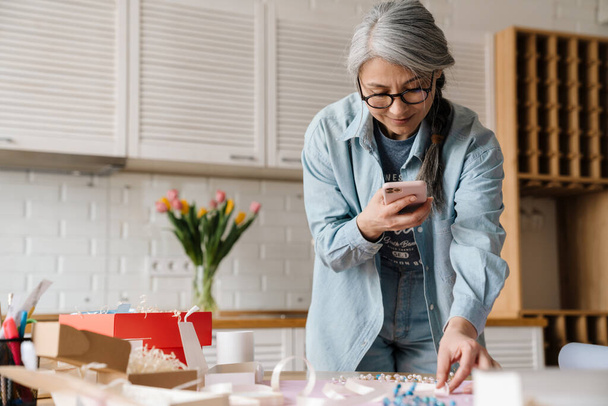 Mature grey woman in eyeglasses taking photo of jewelery on her cellphone at home - Photo, image