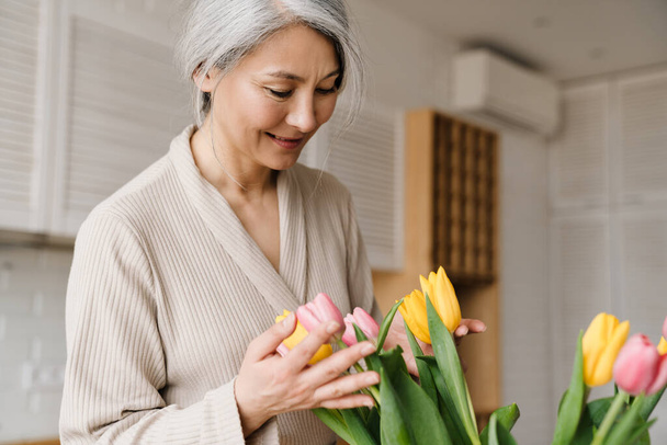 Mature grey woman smiling while holding tulips in kitchen at home - Фото, изображение