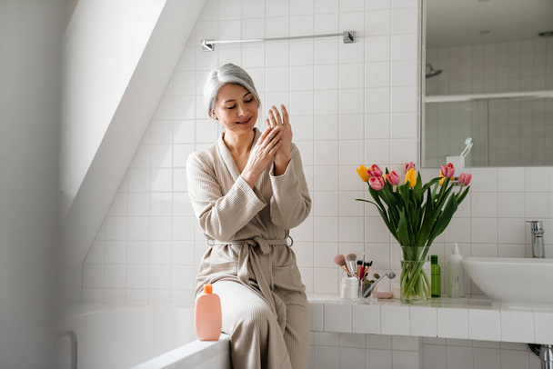 Mature smiling woman applying cream while sitting in bathroom at home - Fotoğraf, Görsel