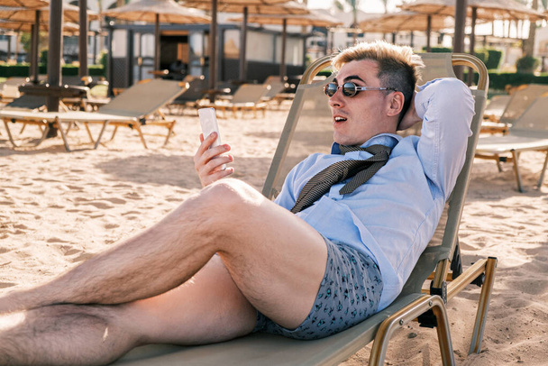 Έκπληκτος χαλαρός επιχειρηματίας ξαπλωμένος σε μια ξαπλώστρα στην παραλία κοιτάζοντας το τηλέφωνο, φοράει πουκάμισο, γραβάτα και κιλότα, ρίχνοντας το ένα χέρι πίσω από το κεφάλι του, με σταυρωμένα τα πόδια. Ο άνθρωπος έχει ιδρωμένη μασχάλη - Φωτογραφία, εικόνα
