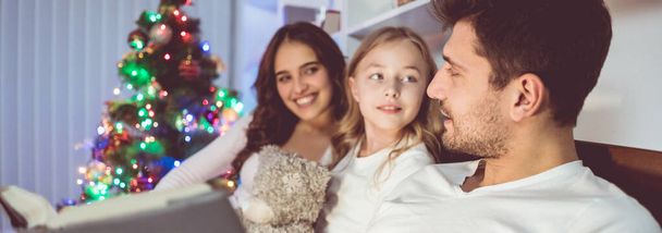 The daughter and parents lay on the bed and read a book near the christmas tree - Photo, image