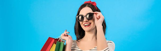 Счастливая женщина в солнечных очках держит цветные мешки на синем фоне - Фото, изображение