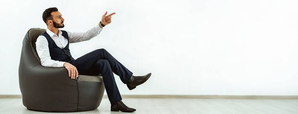 Бізнесмен сидить на мішкуватому стільці на білому тлі стіни і жестикулює
 - Фото, зображення