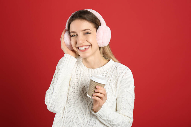 Happy woman with cup of drink wearing warm earmuffs on red background - Photo, Image