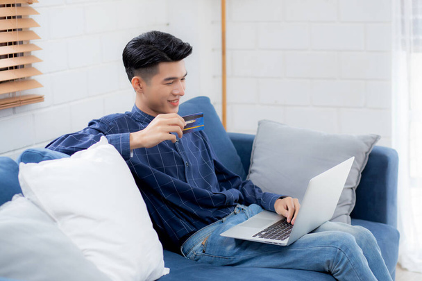 若いアジアの男性は、インターネットへのクレジットカードの購入、ソファでのビジネスと幸せな男性の支払い、購入と支払い、ビジネスコンセプトとラップトップコンピュータのオンラインショッピングを使用してソファに座っている. - 写真・画像