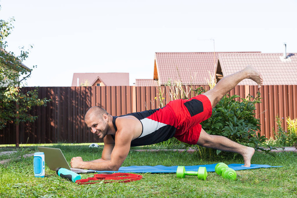 The young man goes in for sports outside. Cheerful sportsman with black hair makes the lunges on lawn in garden, next to there is a laptop with online training - Photo, Image
