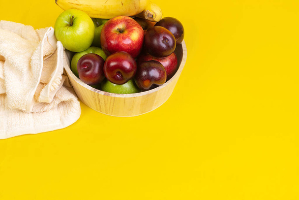 A high angle shot of fresh ripe fruits in a wooden vase on a yellow surface with a towel on it - Photo, Image
