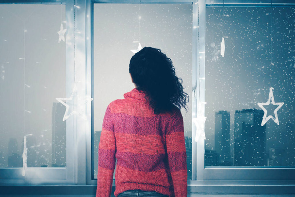 Rear view of young woman wearing warm clothes while standing near the window and enjoying snowfall scenery at winter time. Shot in the apartment - Photo, Image