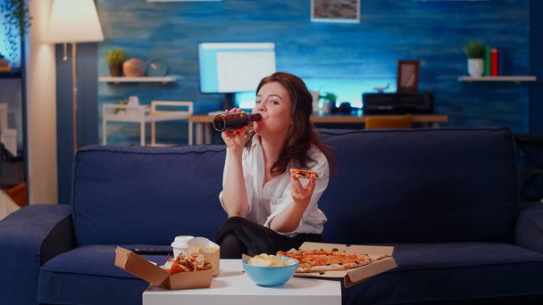 Person eating slice of pizza on couch and looking at camera - Фото, изображение