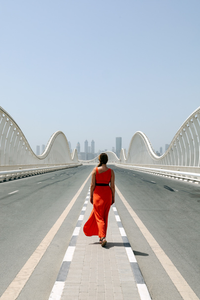 Meydan Köprüsü 'nde boş bir yolda yürüyen turuncu elbiseli bir kadın. Dubai' de şehir manzaralı. - Fotoğraf, Görsel