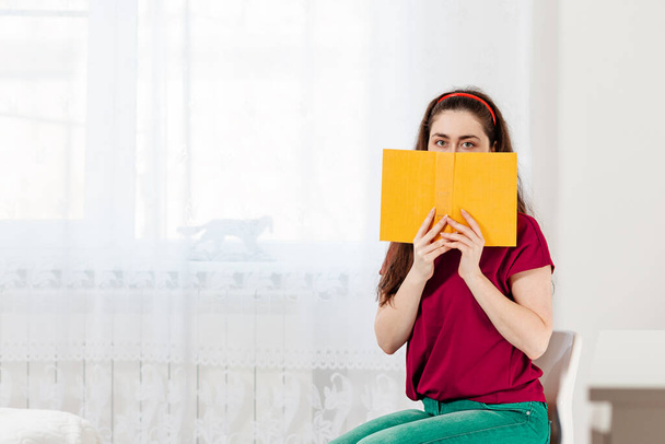 Una donna è seduta sulla sedia e si copre il viso con un libro giallo. Una stanza luminosa sullo sfondo. Concetto di istruzione e lettura. - Foto, immagini