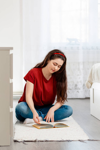 A young, pretty Caucasian woman is sitting cross-legged on the floor, leafing through a book with a smile. Copy space. In the background-the white interior of the room. Concept of education and reading books. - Foto, Bild