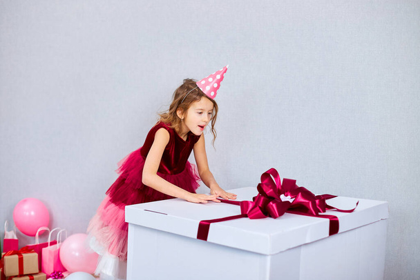 Pembe elbiseli ve şapkalı küçük neşeli kız doğum günü partisinde balonlarla hediye kutusunu açıyor, mutlu yıllar. Kutlama. - Fotoğraf, Görsel