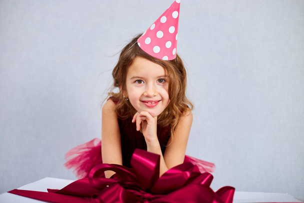 Portrait of joyful little girl in pink dress and hat put her elbows on big present gift box, at home birthday party streamers, Happy birthday. Celebrating - Fotografie, Obrázek
