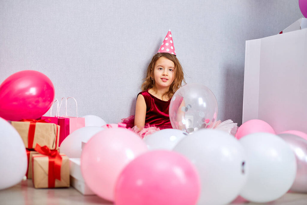 Little girl in pink dress and birthday hat sitting at the floor with many present gift box, balloons at home birthday party streamers, Happy birthday. Celebrating - Photo, Image