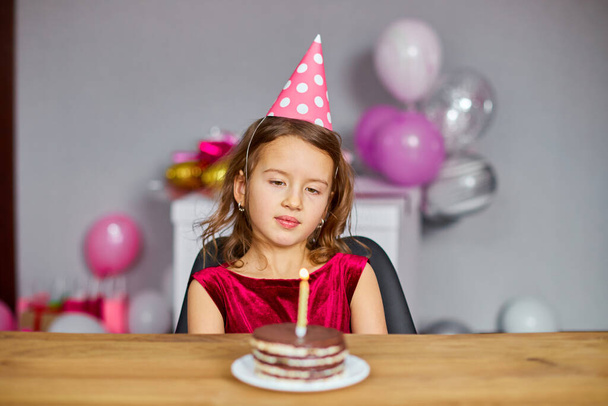 Ein kleines Mädchen trägt einen Geburtstagshut und wünscht sich eine Geburtstagstorte mit leuchtenden Kerzen für ein Party-Konzept. - Foto, Bild