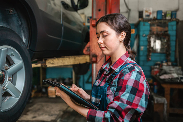 Portrait of a young female mechanic in uniform using a tablet. In the background is an auto repair shop. - Photo, Image