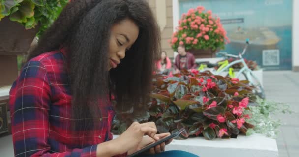Спокійна прогулянка привабливою молодою африканською дівчиною, спілкування, перегляд і смс через планшет, сидячи на відкритому повітрі біля квітів
. - Кадри, відео