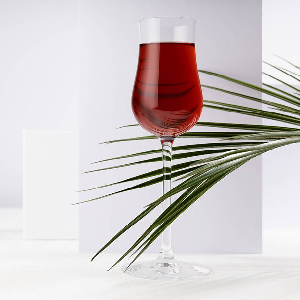 Red porto wine in luxury high glass with green palm leaf in hard light with shadow in soft light white abstract interior with geometric surfaces in minimal style, square. - Fotoğraf, Görsel