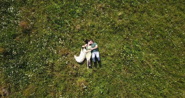 Top portrait of the happy charming couple softly kissing while lying with their dog on the meadow. - Foto, imagen