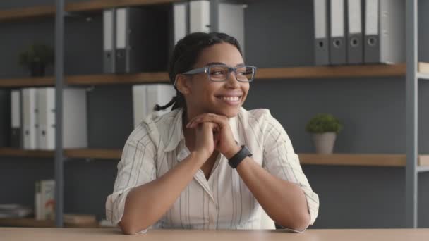 Jeune femme afro-américaine paisible portant des lunettes assis sur le lieu de travail et regardant de côté la fenêtre avec le sourire - Séquence, vidéo