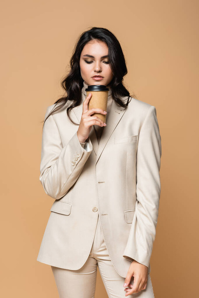 young pretty woman in turtleneck and suit holding paper cup isolated on beige - Fotografie, Obrázek