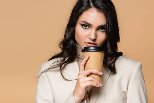 young pretty woman in turtleneck and blazer holding paper cup isolated on beige - Photo, Image