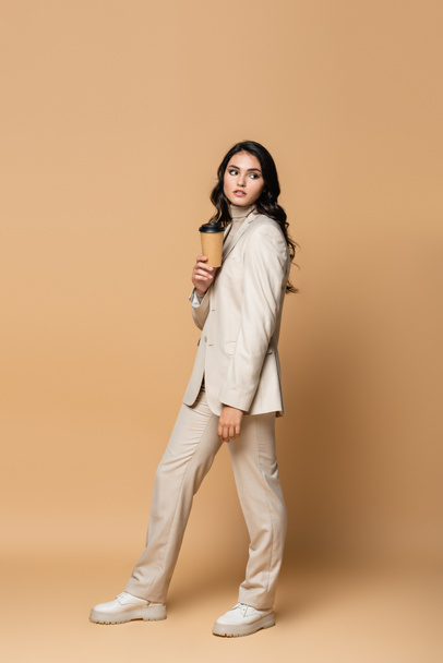 full length of young woman in turtleneck and suit holding coffee to go on beige - Фото, изображение