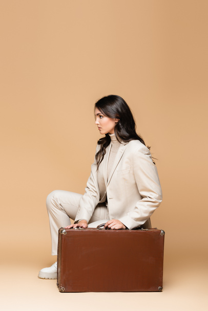 side view of young traveler in suit posing near vintage suitcase on beige  - Valokuva, kuva