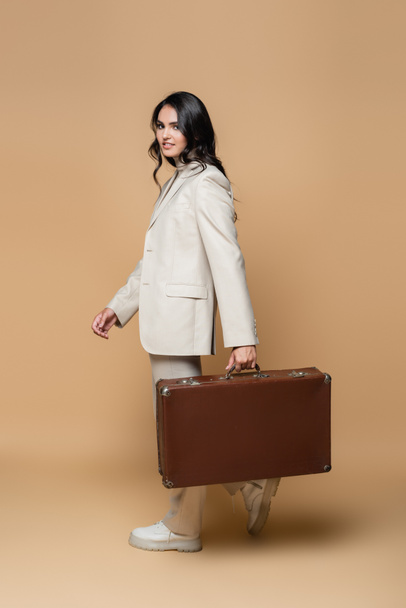 happy young woman in suit carrying vintage suitcase on beige  - Foto, imagen