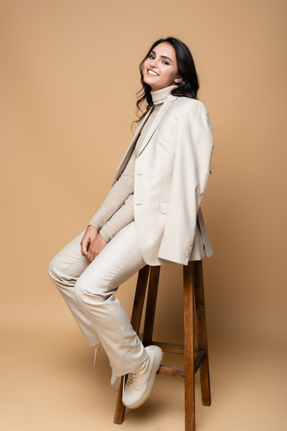 full length of cheerful woman in suit posing while sitting on wooden stool on beige  - Φωτογραφία, εικόνα