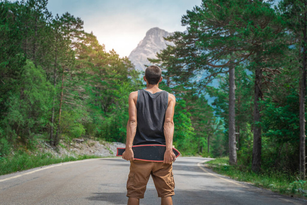 Young hipster holding a cool longboard in his hand. Beautiful concept of travel. Millennial journey of adventure. Skateboard skating ride. Sports in wild nature. - Photo, image