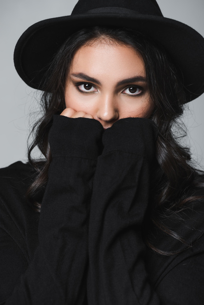 young model in black fedora hat and turtleneck looking at camera while covering mouth isolated on grey - Photo, Image