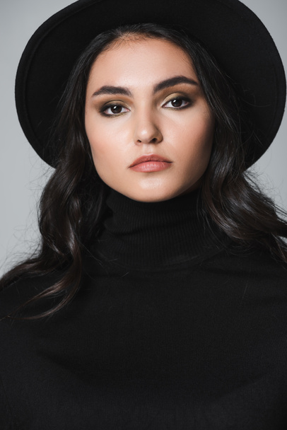 pretty model in black fedora hat and turtleneck looking at camera isolated on grey - Fotó, kép