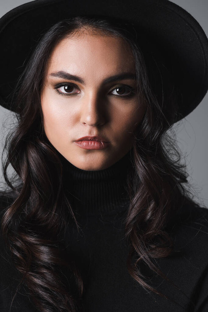 brunette model in black fedora hat and turtleneck looking at camera isolated on grey - Foto, immagini