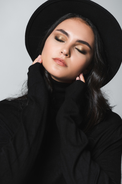 young model with closed eyes in black fedora hat and turtleneck isolated on grey - Foto, afbeelding