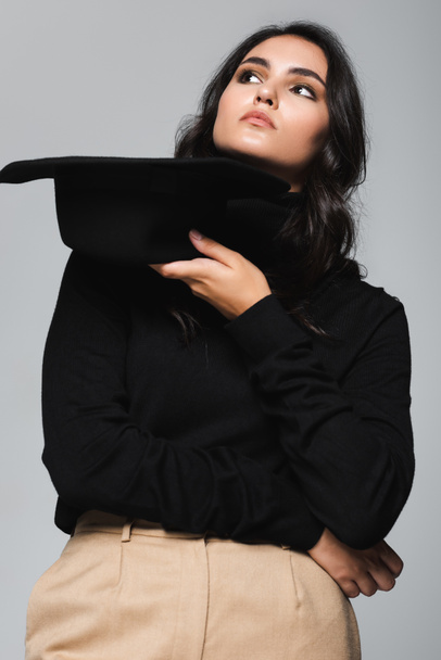 молода модель в черепашці, що тримає чорний капелюх фідори ізольований на сірому
 - Фото, зображення