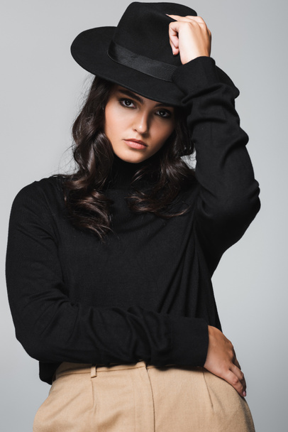 brunette and young model holding fedora hat near head isolated on grey - Фото, зображення