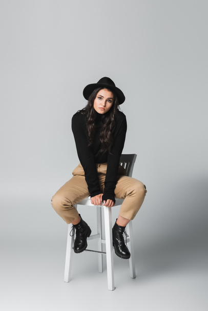full length of young and trendy woman in fedora hat looking at camera while sitting on chair on grey  - 写真・画像