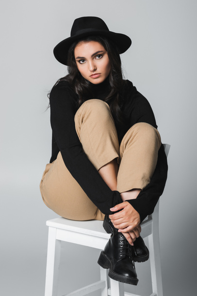 full length of brunette model in fedora hat looking at camera while sitting on chair on grey  - Photo, Image