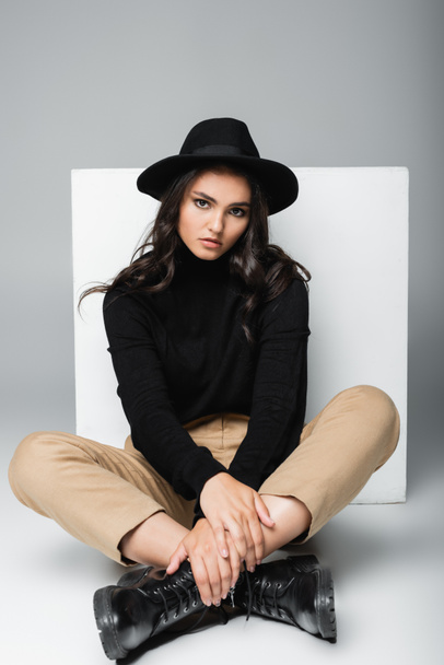 full length of stylish model in fedora hat sitting with crossed legs near white cube on grey  - Foto, afbeelding