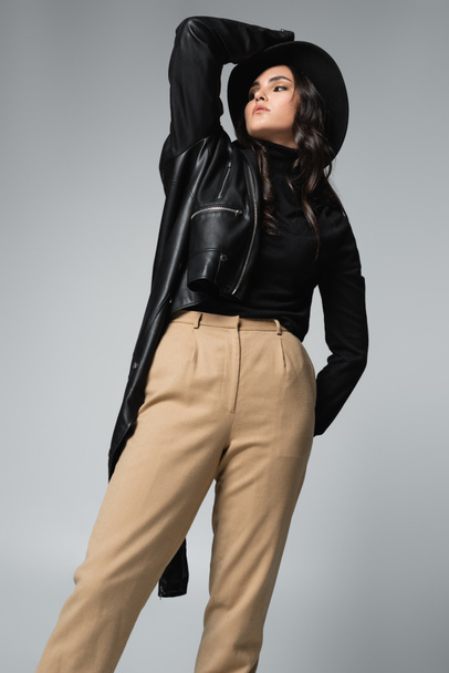 low angle view of trendy woman in fedora hat and black leather jacket posing isolated on grey  - Zdjęcie, obraz