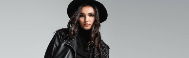 stylish woman in fedora hat and black leather jacket posing isolated on grey, banner  - 写真・画像