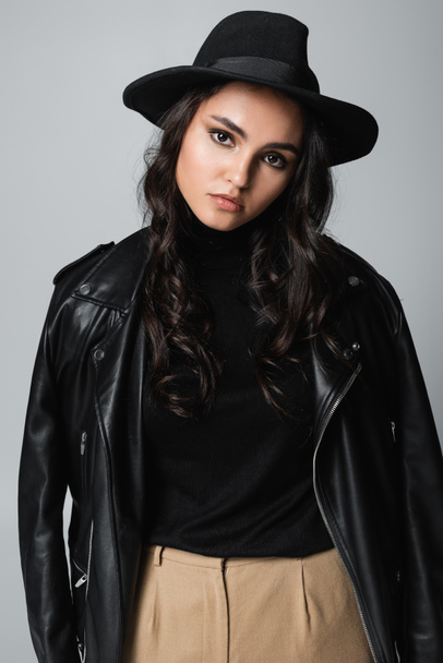 young brunette woman in fedora hat, black turtleneck and trendy leather jacket posing isolated on grey  - Fotografie, Obrázek