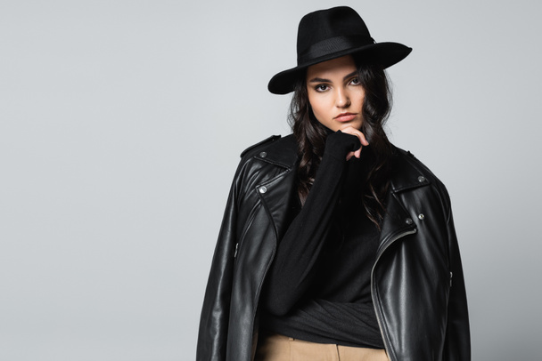 young woman in fedora hat, black turtleneck and trendy leather jacket posing isolated on grey  - Фото, зображення