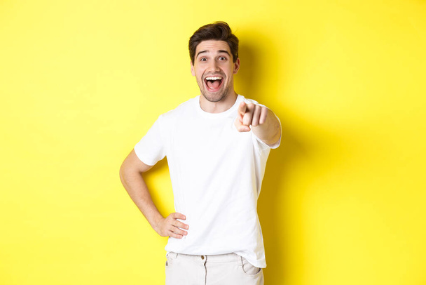 Happy guy pointing finger at camera and laughing, check out something, standing over yellow background - Photo, Image