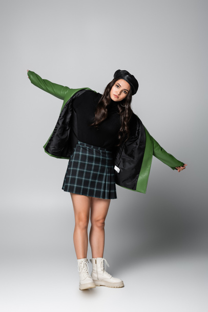 full length of young stylish woman in beret, plaid skirt and green leather jacket posing with outstretched hands on grey - 写真・画像
